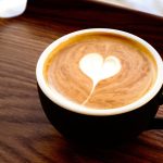 Crna šalica s kremastim cappuccinom i srcolikim latte artom poslužena na drvenom pladnju.