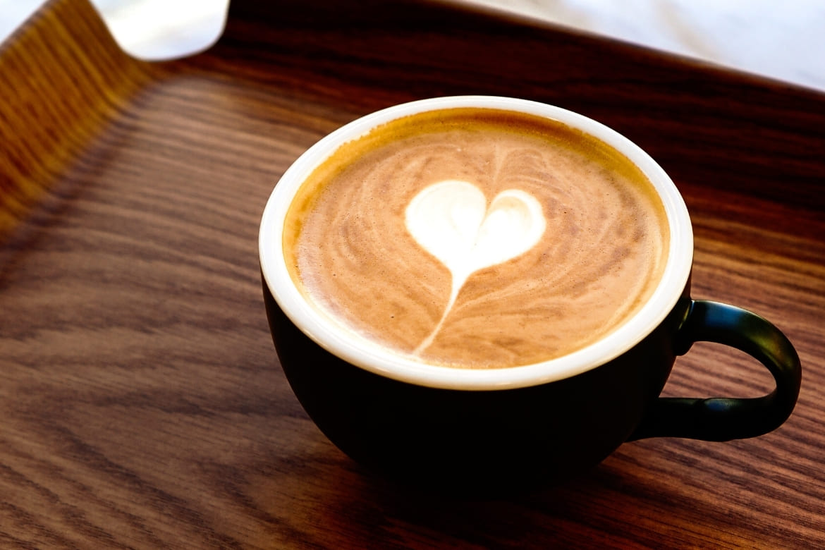 Crna šalica s kremastim cappuccinom i srcolikim latte artom poslužena na drvenom pladnju.