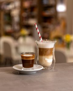 Dvije šalice kave na stolu u kafiću. Lijevo je mala staklena čaša s tamnim espresso macchiatom, a desno visoka staklena čaša s latte macchiatom, s jasnim slojevima mlijeka, kave i pjene.