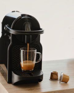 Crni aparat za kavu toči svježi espresso s bogatom pjenom u prozirnu staklenu šalicu uz iskorištenu kapsulu.