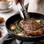 Pečenje sočnog odreska u tavi s ružmarinom i češnjakom, uz vidljiva zrna kave koja sugeriraju marinadu od kave.