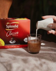 Ulijevanje svježeg mlijeka iz vrča u čašu s espressom pokraj kutije Splendid Classico kompatibilnih kapsula.